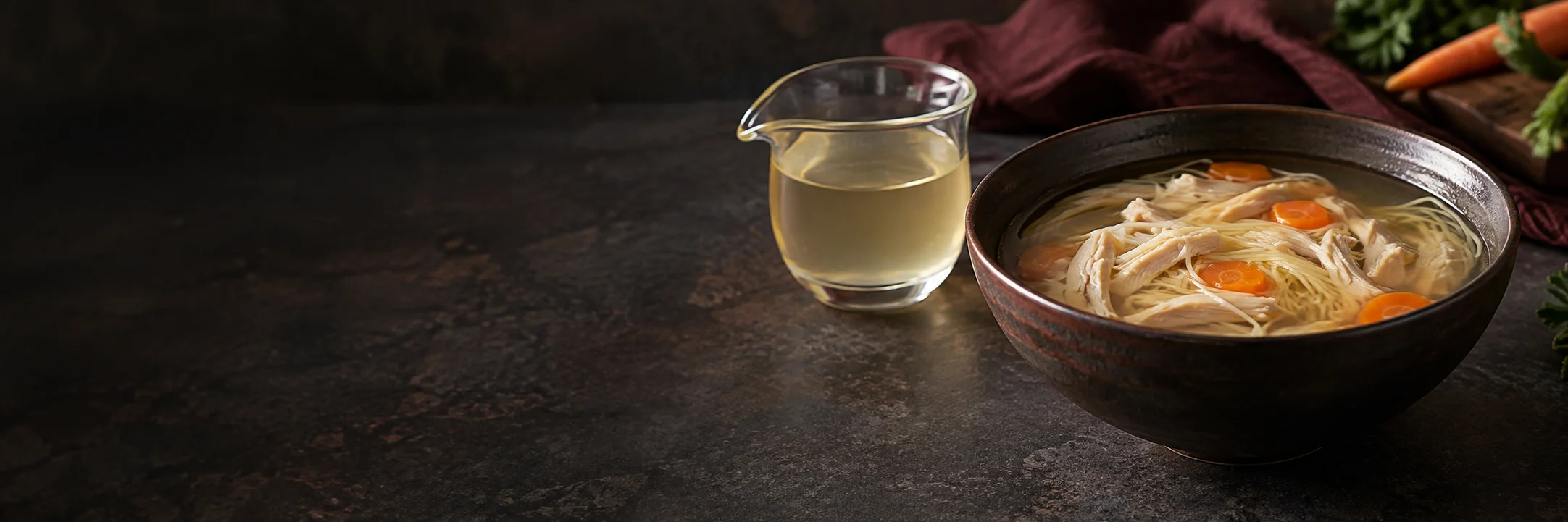 A glass vessel of pale clear broth and a bowl of noodle soup with white chicken pieces and carrots illustrating soups broths stocks product-format context.