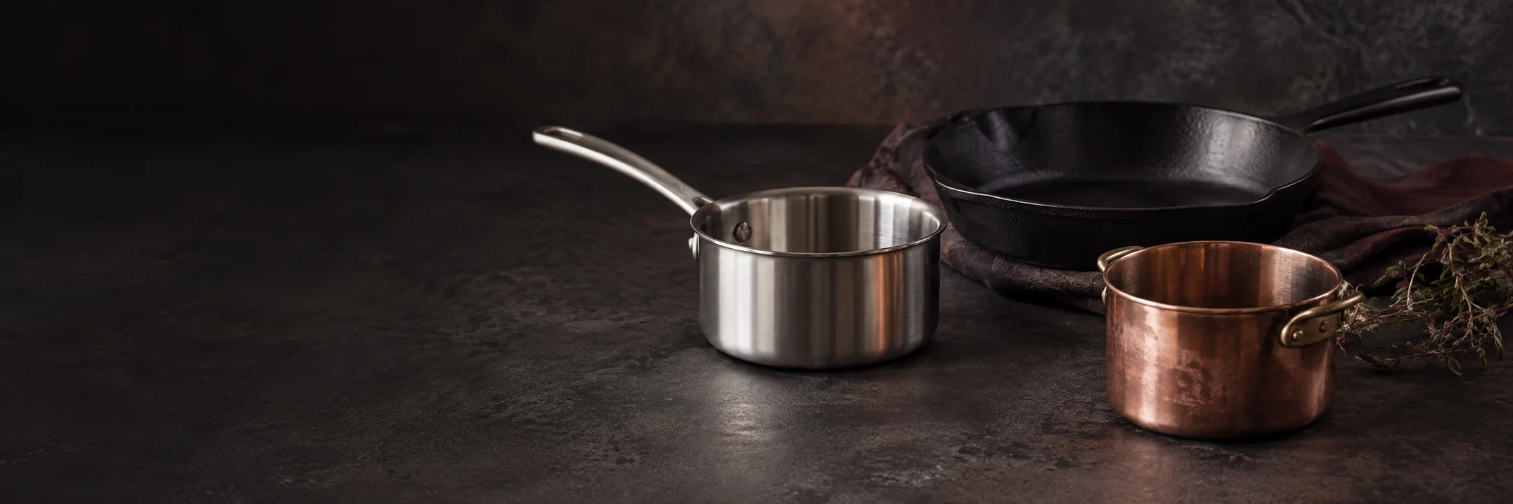 A stainless-steel saucepan, cast-iron skillet and copper-colored pot illustrating china gb 4806 9 2023 food contact metal research context.