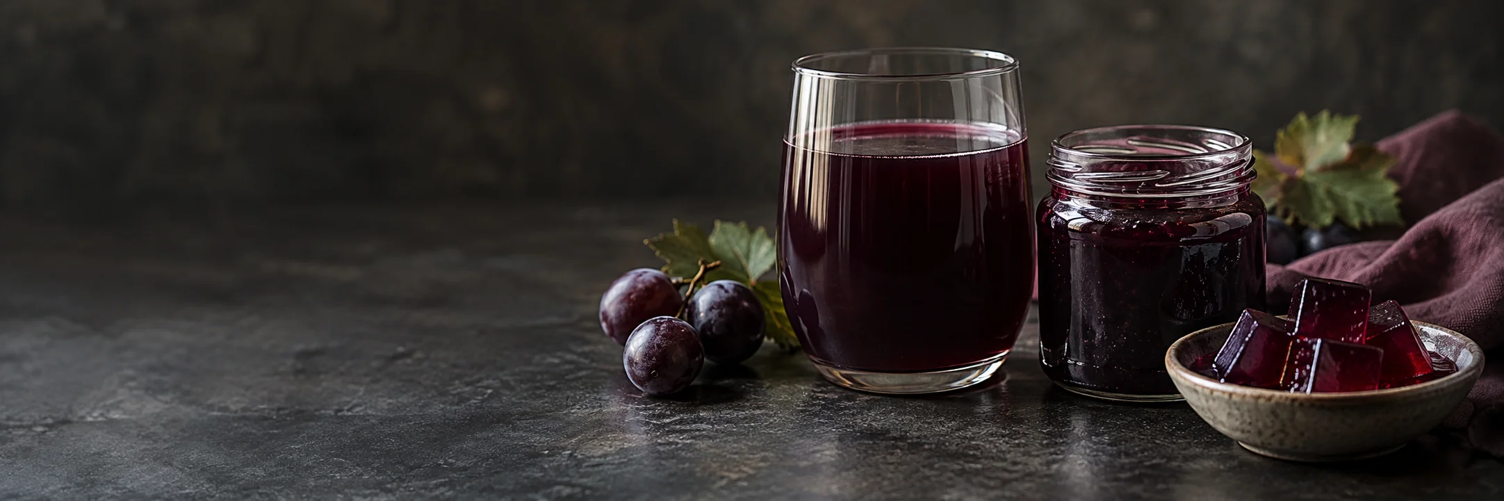 A glass of dark purple juice, an unmarked jelly jar, translucent dessert-gel cubes and dark grapes illustrating grape jelly ingredient context.