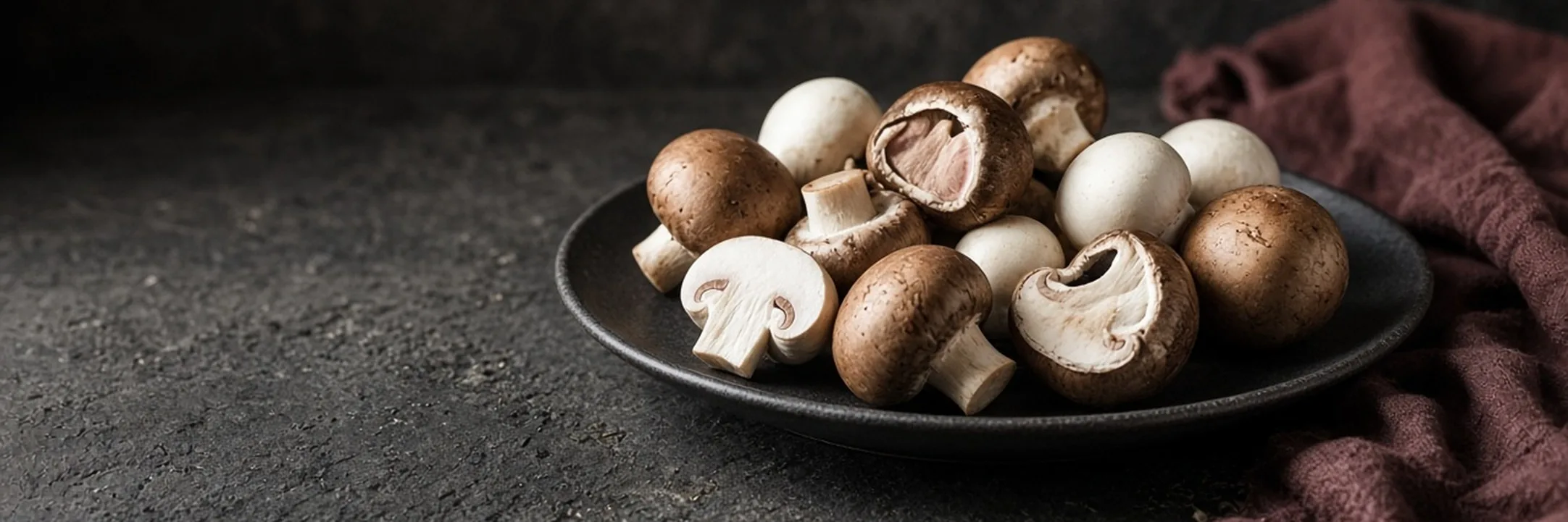 Whole and halved brown cremini and white button mushrooms on a dark ceramic plate beside a burgundy cloth.