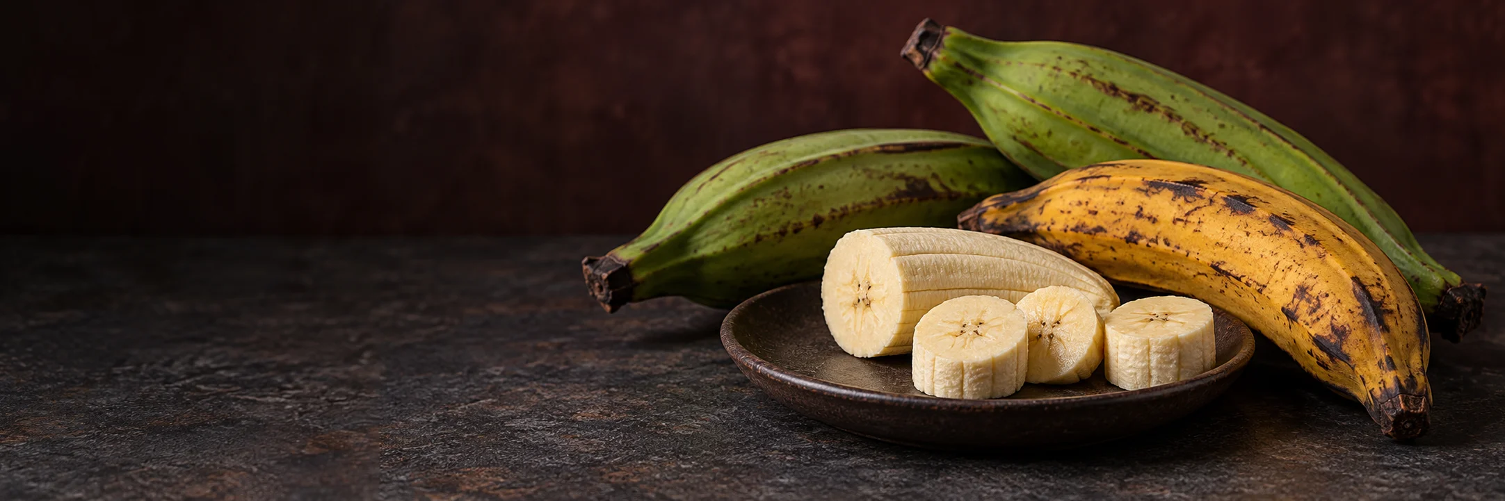 Green and ripe plantains beside a plate of peeled plantain slices illustrating plantain ingredient context.